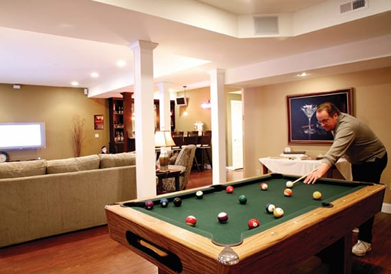Basement remodel with wood floors, beige walls, pool table in foreground with person playing pool on right side, and back of a long couch and view of TV screen in background/to the left.