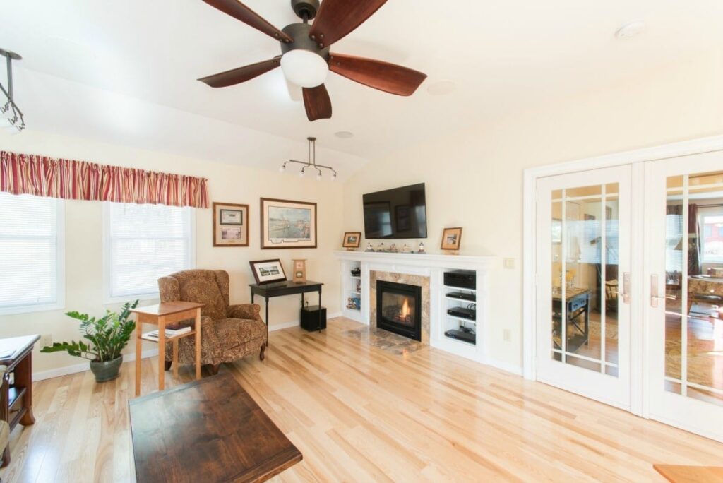 Living area home addition with light wood floors, gas fireplace in far corner with white built-in shelving on either side, ceiling fan, white walls, and double glass doors leading to rest of home.