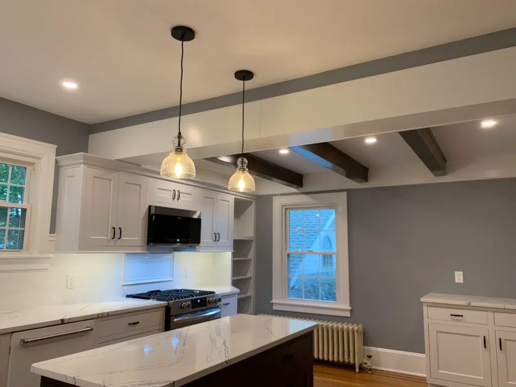 Updated kitchen with island. Wood floors, gray walls, wood beams in ceiling, white cabinets against wall, black cabinets for island, white stone countertop and white tile backsplash.
