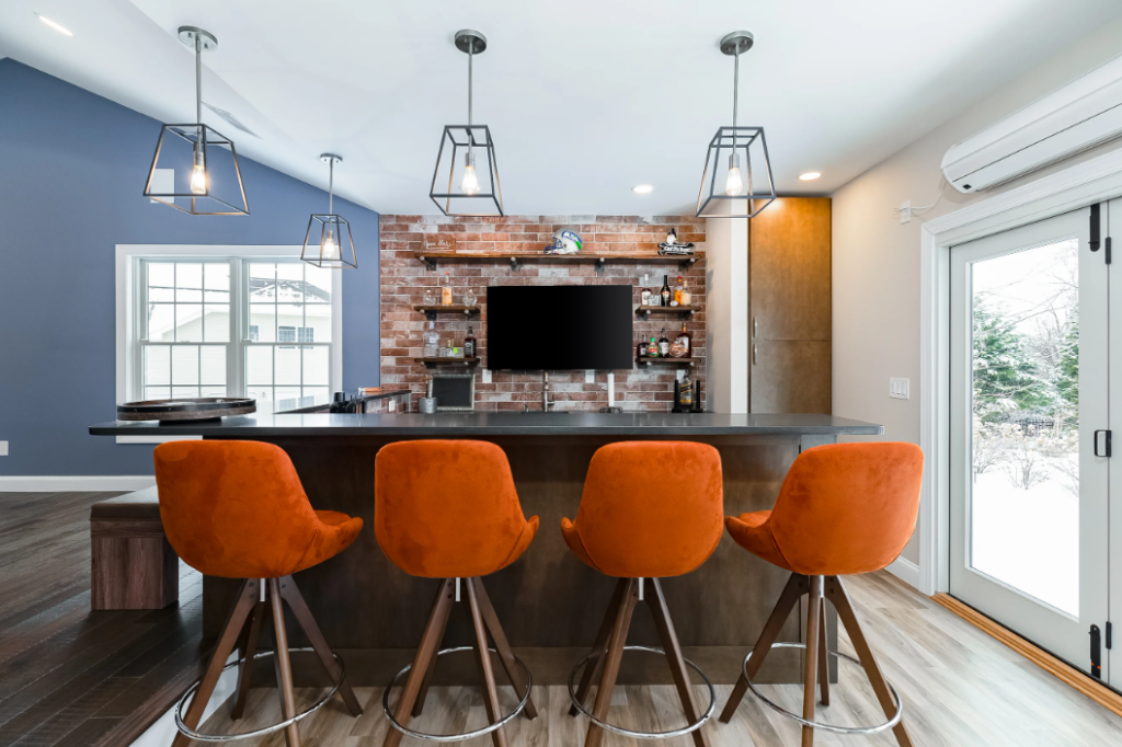 Remodeled home bar area with wood floors, peninsula counters with dark cabinets and black countertop, orange stool seating in foreground, brick backsplash on far wall with open woodgrain shelving around a wall-mounted TV.