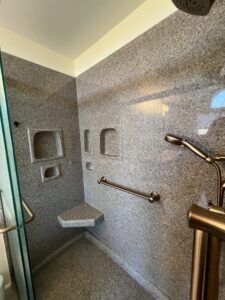 Bathroom remodel with brown stone-look shower tile, walk-in glass-enclosed shower with grab bars and corner seat.