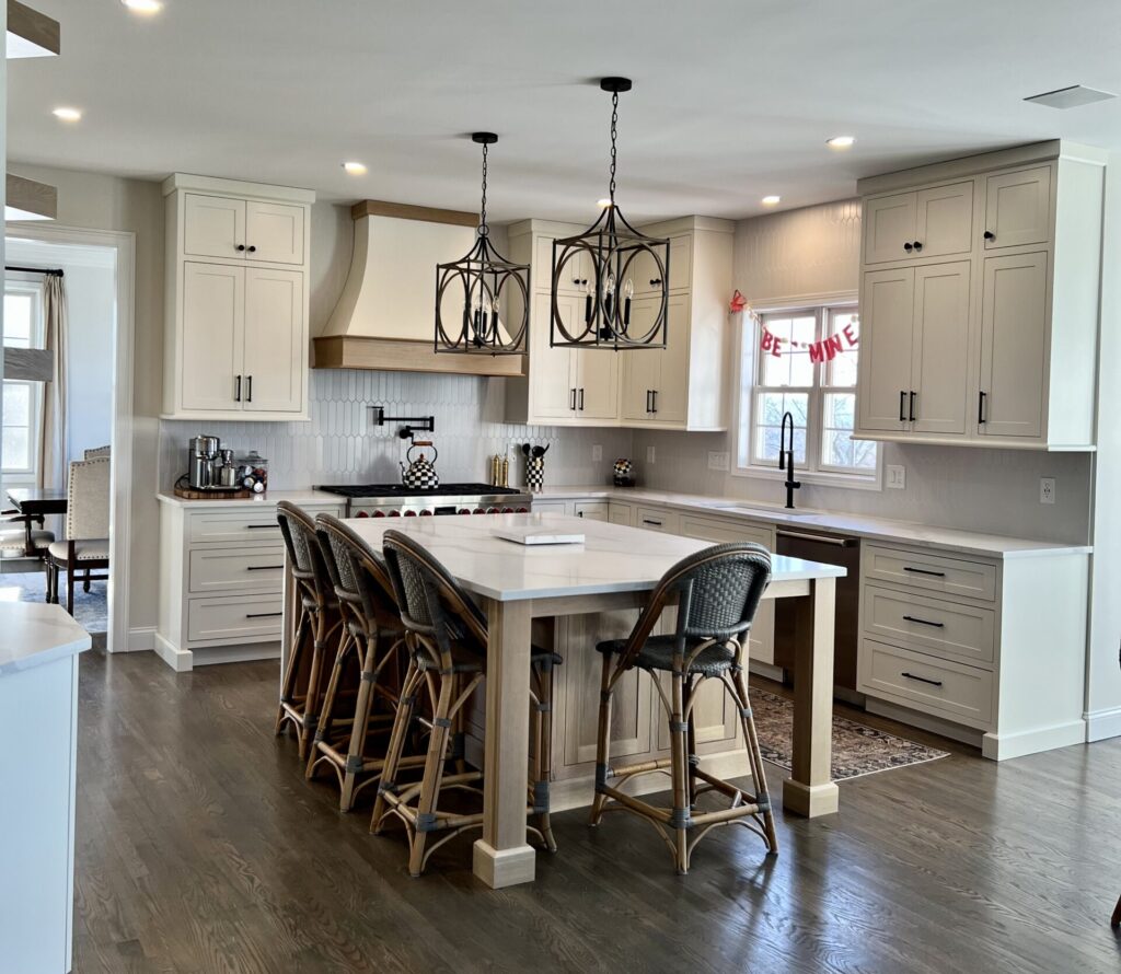 Remodeled kitchen with wood floors, white cabinets, square island with seating on two sides, white stone countertops, white vertical tile backsplash, recessed and pendant lighting.